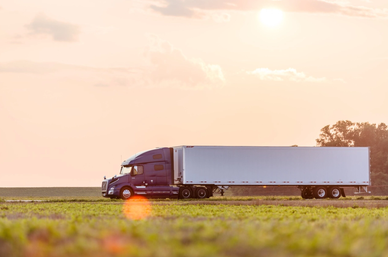 A semi-truck with a white refrigerated trailer drives through a rural landscape at sunset, with the sun casting a warm glow over the open fields.